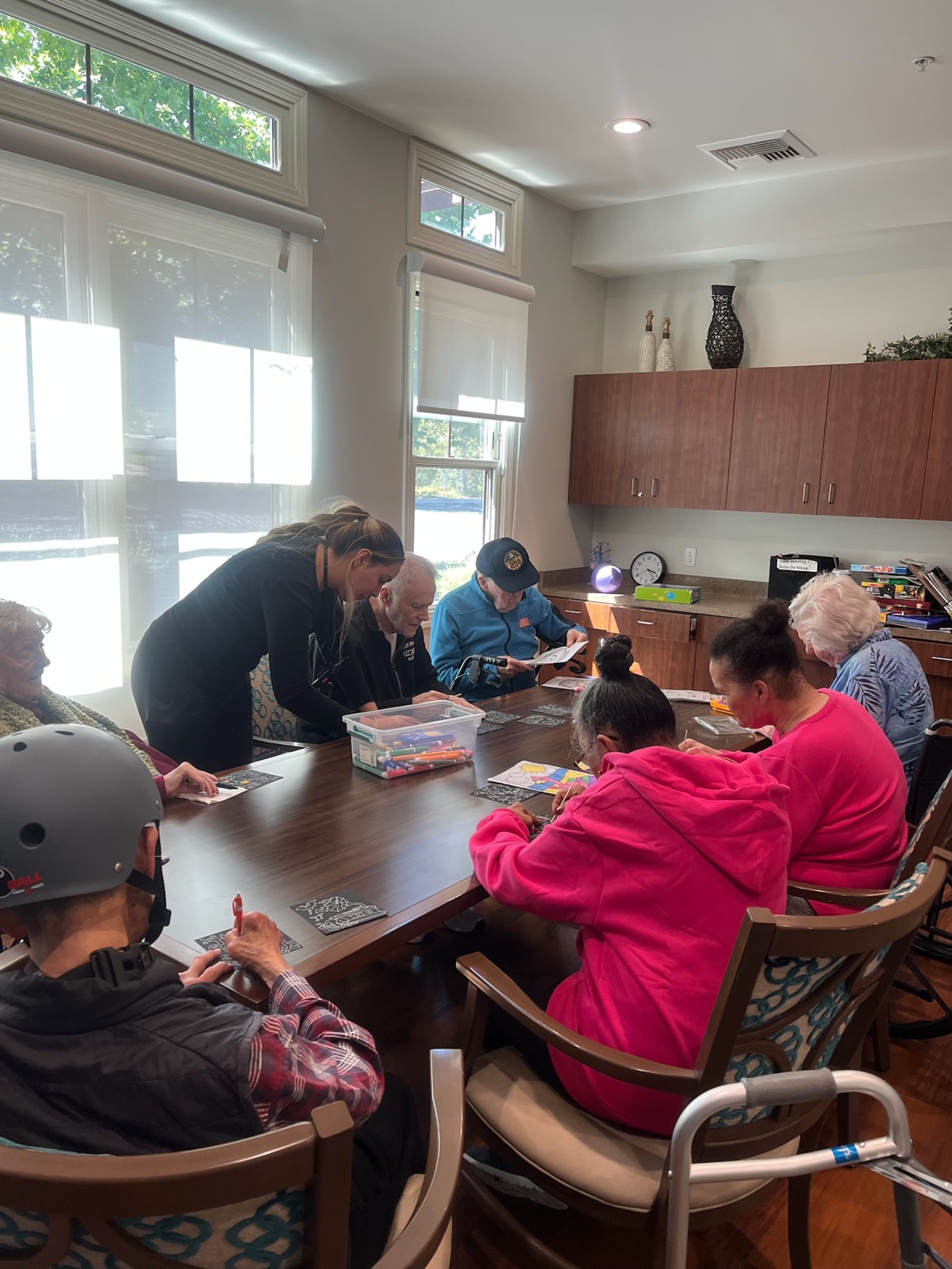 A facilitator leaning in to help residents with scratch-art cards at a long table